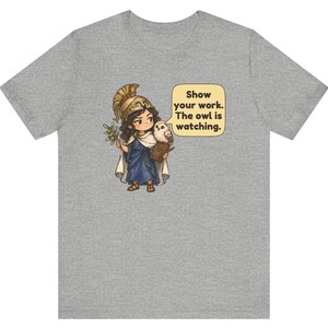 Puede incluir: Una camiseta gris jaspeado con una ilustraci&oacute;n de un personaje con casco dorado y t&uacute;nica azul que sostiene una rama de olivo y un libro. Un peque&ntilde;o b&uacute;ho est&aacute; sobre el hombro junto a un globo de texto que dice, Show your work. The owl is watching.