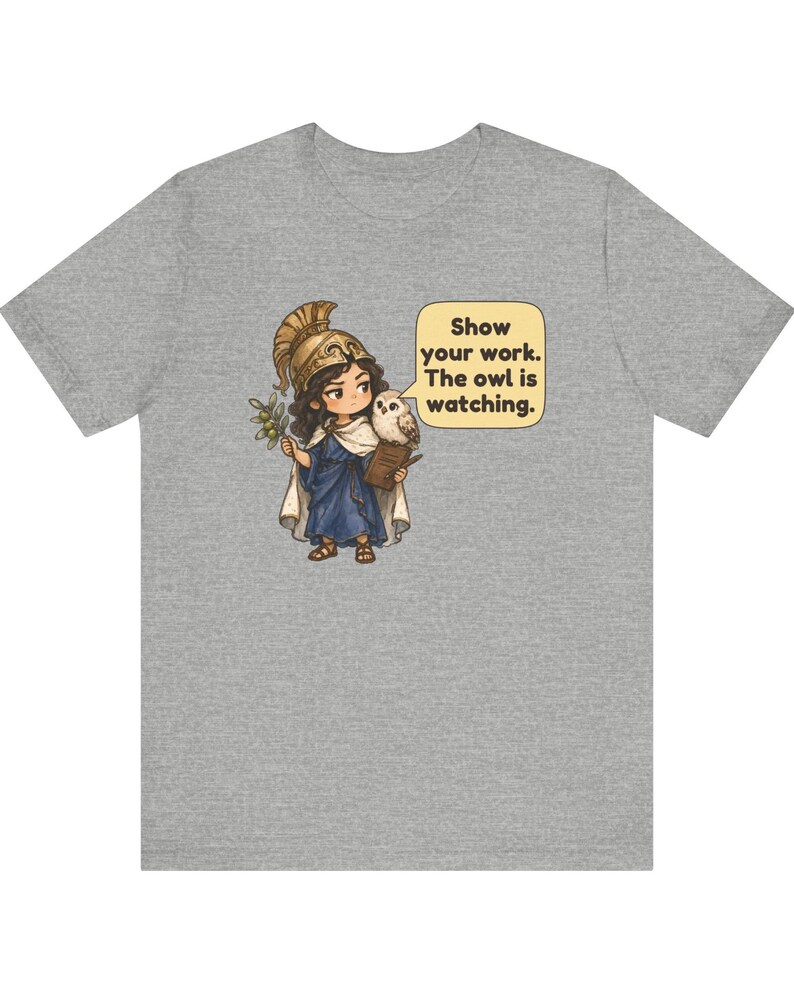 Puede incluir: Una camiseta gris jaspeado con una ilustraci&oacute;n de un personaje con casco dorado y t&uacute;nica azul que sostiene una rama de olivo y un libro. Un peque&ntilde;o b&uacute;ho est&aacute; sobre el hombro junto a un globo de texto que dice, Show your work. The owl is watching.