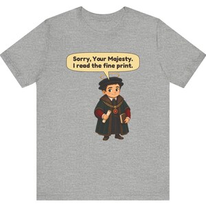 May include: A heather gray t-shirt featuring a cartoon illustration of a person wearing a dark robe, a gold chain, and a halo. The person holds a scroll and a book. A speech bubble above contains the text: Sorry, Your Majesty. I read the fine print.