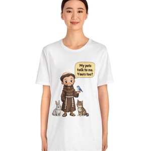 May include: A white t-shirt featuring a cartoon illustration of a monk in a brown robe standing with a gray rabbit, a brown wolf, and a blue bird. A yellow speech bubble above the monk contains the text: My pets talk to me. Yours too?