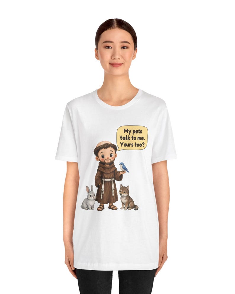 May include: A white t-shirt featuring a cartoon illustration of a monk in a brown robe standing with a gray rabbit, a brown wolf, and a blue bird. A yellow speech bubble above the monk contains the text: My pets talk to me. Yours too?