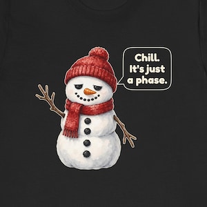 Pode incluir: Uma t-shirt preta com um gr&aacute;fico de um boneco de neve branco a usar um gorro e um cachecol de malha vermelhos. O boneco de neve tem bra&ccedil;os de pau, nariz de cenoura e bot&otilde;es de carv&atilde;o. Um bal&atilde;o de fala cont&eacute;m o texto, Chill. It's just a phase.