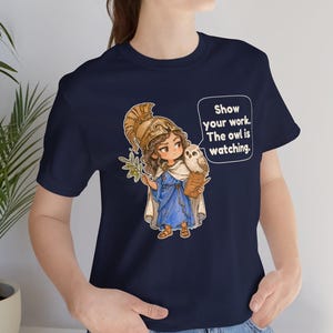 Puede incluir: Una camiseta azul marino con un gr&aacute;fico de un personaje con casco dorado y t&uacute;nica azul, sosteniendo una rama de olivo y un portapapeles con un b&uacute;ho. Un globo de texto contiene el mensaje: Show your work. The owl is watching.