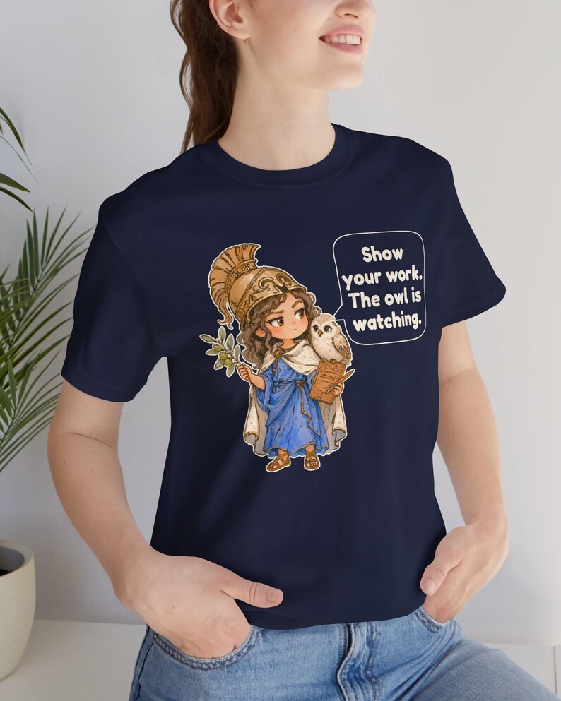 Puede incluir: Una camiseta azul marino con un gr&aacute;fico de un personaje con casco dorado y t&uacute;nica azul, sosteniendo una rama de olivo y un portapapeles con un b&uacute;ho. Un globo de texto contiene el mensaje: Show your work. The owl is watching.