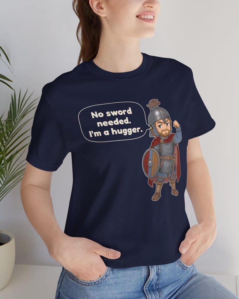 May include: A navy blue t-shirt featuring a cartoon illustration of a warrior in chainmail armor, a red cape, and a helmet, holding a round shield. A speech bubble next to the character contains the text: No sword needed. I'm a hugger.
