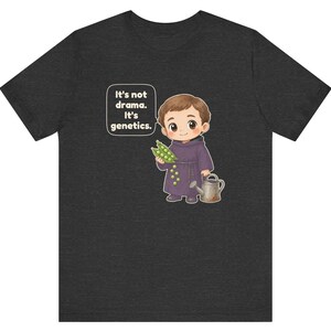 May include: A dark heather gray t-shirt featuring a cartoon illustration of a monk holding green pea pods and a watering can. A speech bubble next to the character contains the text: It's not drama. It's genetics. The design is centered on the chest.
