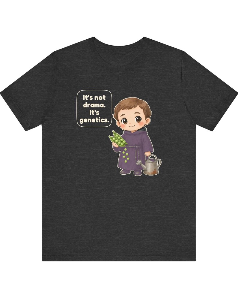 May include: A dark heather gray t-shirt featuring a cartoon illustration of a monk holding green pea pods and a watering can. A speech bubble next to the character contains the text: It's not drama. It's genetics. The design is centered on the chest.
