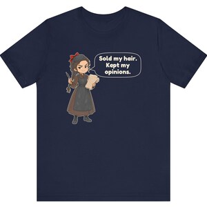 May include: A navy blue short-sleeve t-shirt featuring a cartoon illustration of a person with a red hair bow, brown dress, and apron. The character holds a quill and paper with a speech bubble that reads, Sold my hair. Kept my opinions.