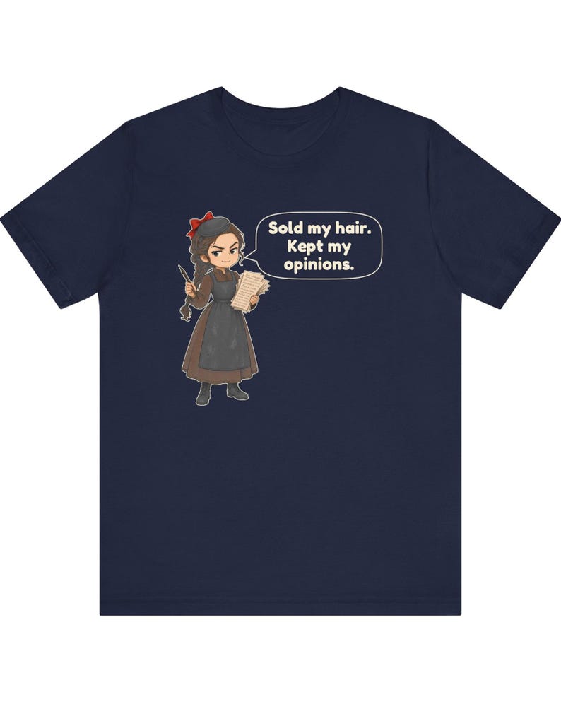 May include: A navy blue short-sleeve t-shirt featuring a cartoon illustration of a person with a red hair bow, brown dress, and apron. The character holds a quill and paper with a speech bubble that reads, Sold my hair. Kept my opinions.