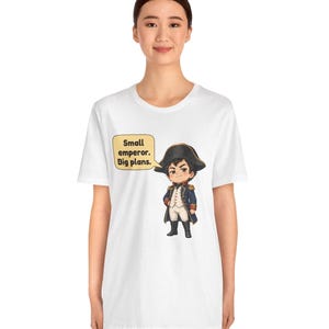 May include: A white short-sleeve t-shirt featuring a graphic of a cartoon character dressed in a historical military uniform with a bicorne hat. A speech bubble next to the character contains the text, Small emperor. Big plans.
