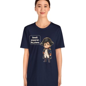 May include: A navy blue short-sleeve t-shirt featuring a cartoon illustration of a historical figure in a military uniform and bicorne hat. A speech bubble next to the character contains the text: Small emperor. Big plans.