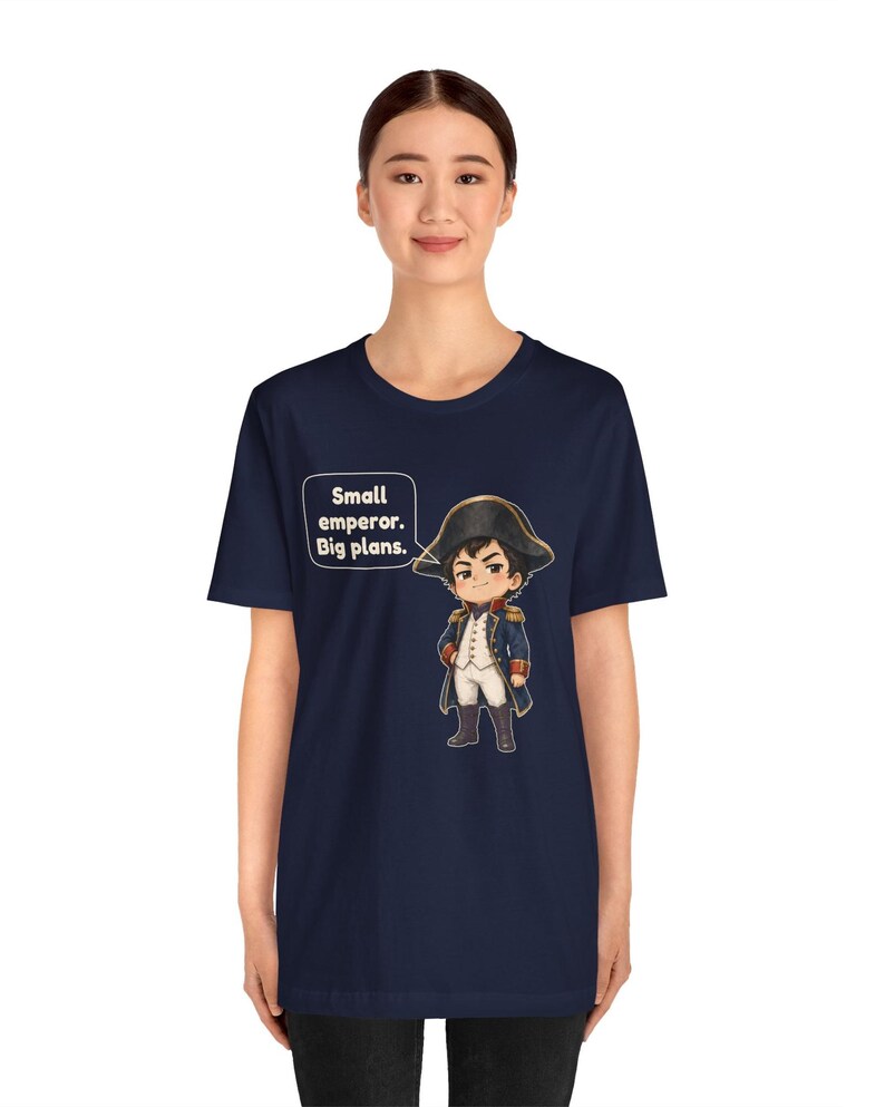 May include: A navy blue short-sleeve t-shirt featuring a cartoon illustration of a historical figure in a military uniform and bicorne hat. A speech bubble next to the character contains the text: Small emperor. Big plans.