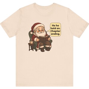 Op de afbeelding: Een lichtbeige t-shirt met een afbeelding van de Kerstman die in een groene fauteuil zit, een boek leest en een dampende mok op een bijzettafeltje heeft staan. Een tekstballon bevat de tekst: Ho ho hold on. Chapter ending.