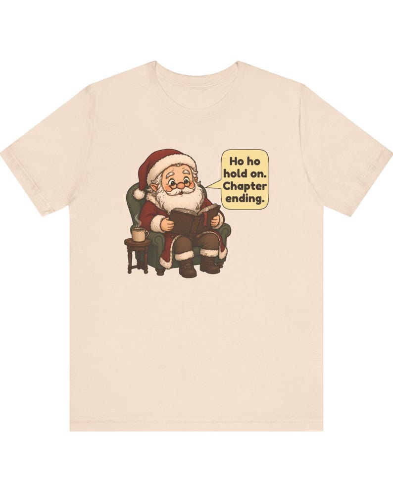 Op de afbeelding: Een lichtbeige t-shirt met een afbeelding van de Kerstman die in een groene fauteuil zit, een boek leest en een dampende mok op een bijzettafeltje heeft staan. Een tekstballon bevat de tekst: Ho ho hold on. Chapter ending.