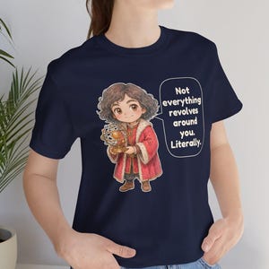 May include: A navy blue t-shirt featuring a cartoon character with curly brown hair wearing a red coat with fur trim, holding a model of the solar system. A speech bubble contains the text: Not everything revolves around you. Literally.
