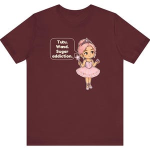 May include: A maroon short-sleeve t-shirt featuring a graphic of a cartoon ballerina with pink hair, a tiara, and a pink tutu. The character holds a star wand and a cupcake. A speech bubble contains the text: Tutu. Wand. Sugar addiction.