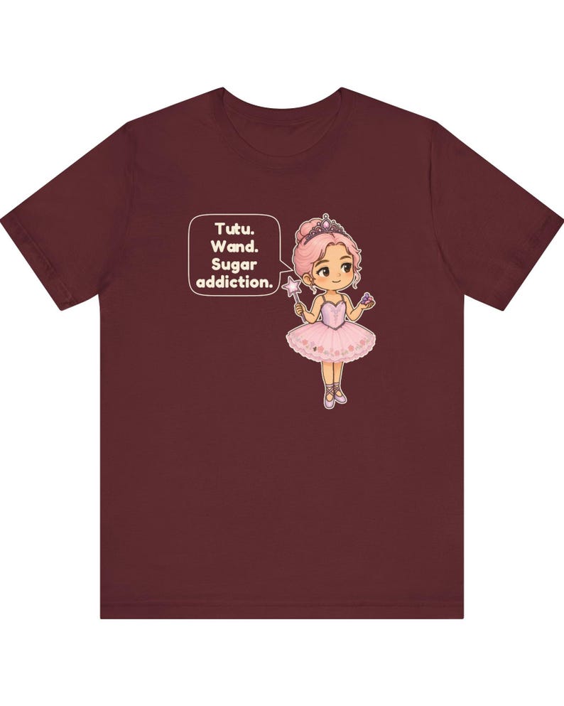 May include: A maroon short-sleeve t-shirt featuring a graphic of a cartoon ballerina with pink hair, a tiara, and a pink tutu. The character holds a star wand and a cupcake. A speech bubble contains the text: Tutu. Wand. Sugar addiction.