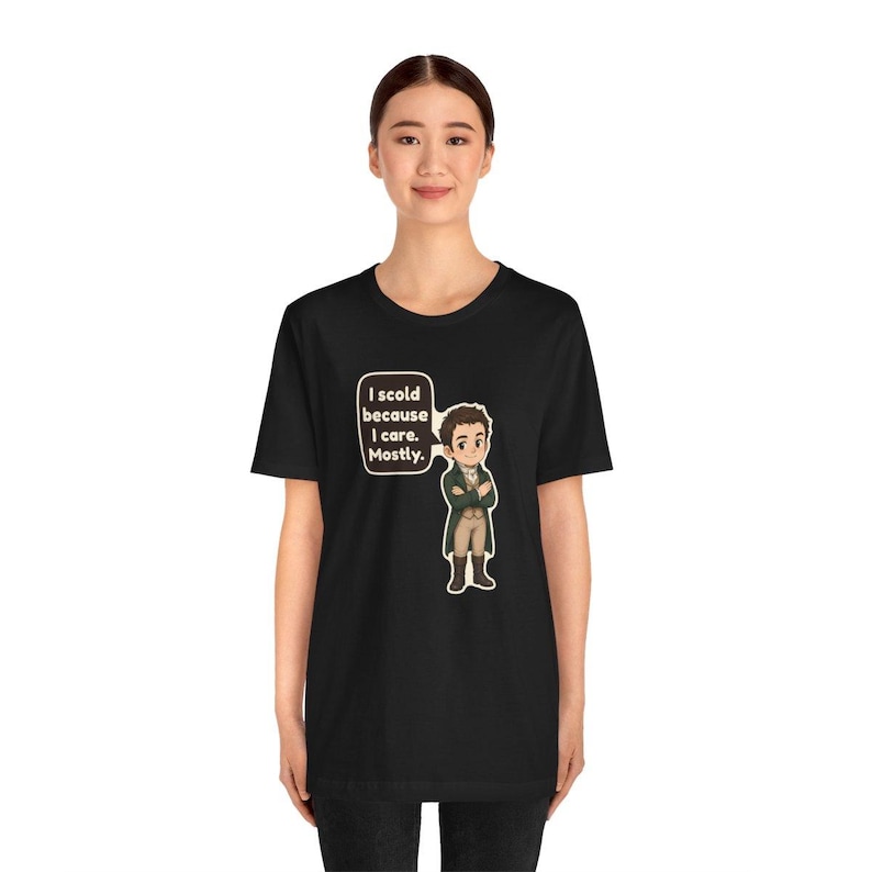 May include: A black t-shirt featuring a cartoon illustration of a person in a green coat and tan pants with arms crossed. A speech bubble next to the character contains the text: I scold because I care. Mostly.