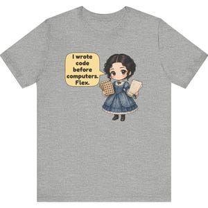 May include: A heather gray t-shirt featuring a cartoon illustration of a person in a blue dress holding punch cards and papers. A yellow speech bubble contains the text: I wrote code before computers. Flex.