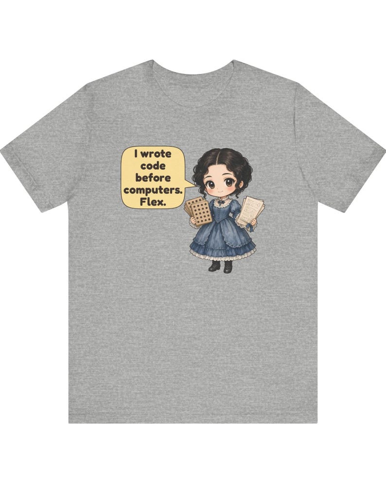 May include: A heather gray t-shirt featuring a cartoon illustration of a person in a blue dress holding punch cards and papers. A yellow speech bubble contains the text: I wrote code before computers. Flex.
