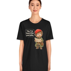 May include: A black short-sleeved t-shirt featuring a graphic of a cartoon character wearing a red cap and green coat, holding a map. A speech bubble above the character contains the text: Yes, I've heard the pool joke.