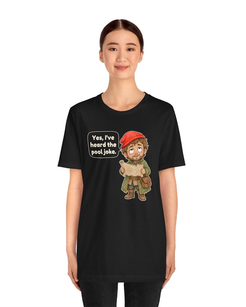 May include: A black short-sleeved t-shirt featuring a graphic of a cartoon character wearing a red cap and green coat, holding a map. A speech bubble above the character contains the text: Yes, I've heard the pool joke.