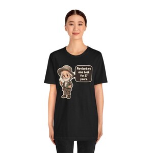 May include: A black short-sleeve t-shirt featuring a cartoon illustration of an elderly man in a brown hat and suit holding a book. A speech bubble next to him contains the text: Revised my one book for 37 years.