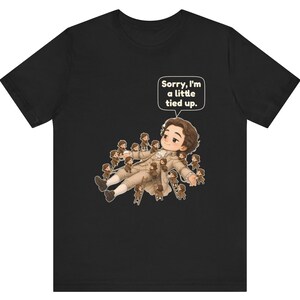 Op de afbeelding: Een zwart t-shirt met korte mouwen en een afbeelding van een groot persoon die ligt, omringd door vele kleine figuren op ladders. Een tekstballon boven de grote figuur bevat de tekst: "Sorry, I'm a little tied up."