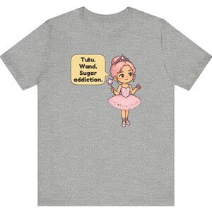May include: A heather gray t-shirt featuring a cartoon illustration of a character with pink hair wearing a pink tutu, a tiara, and ballet slippers. The character holds a star-tipped wand and a cupcake, with a speech bubble saying Tutu. Wand. Sugar addiction.