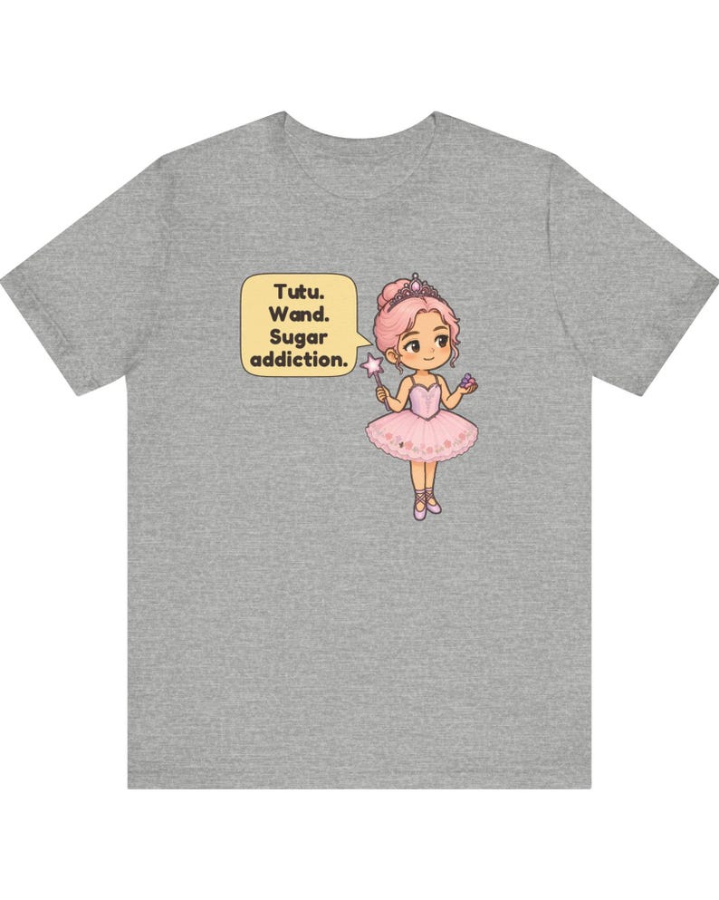 May include: A heather gray t-shirt featuring a cartoon illustration of a character with pink hair wearing a pink tutu, a tiara, and ballet slippers. The character holds a star-tipped wand and a cupcake, with a speech bubble saying Tutu. Wand. Sugar addiction.