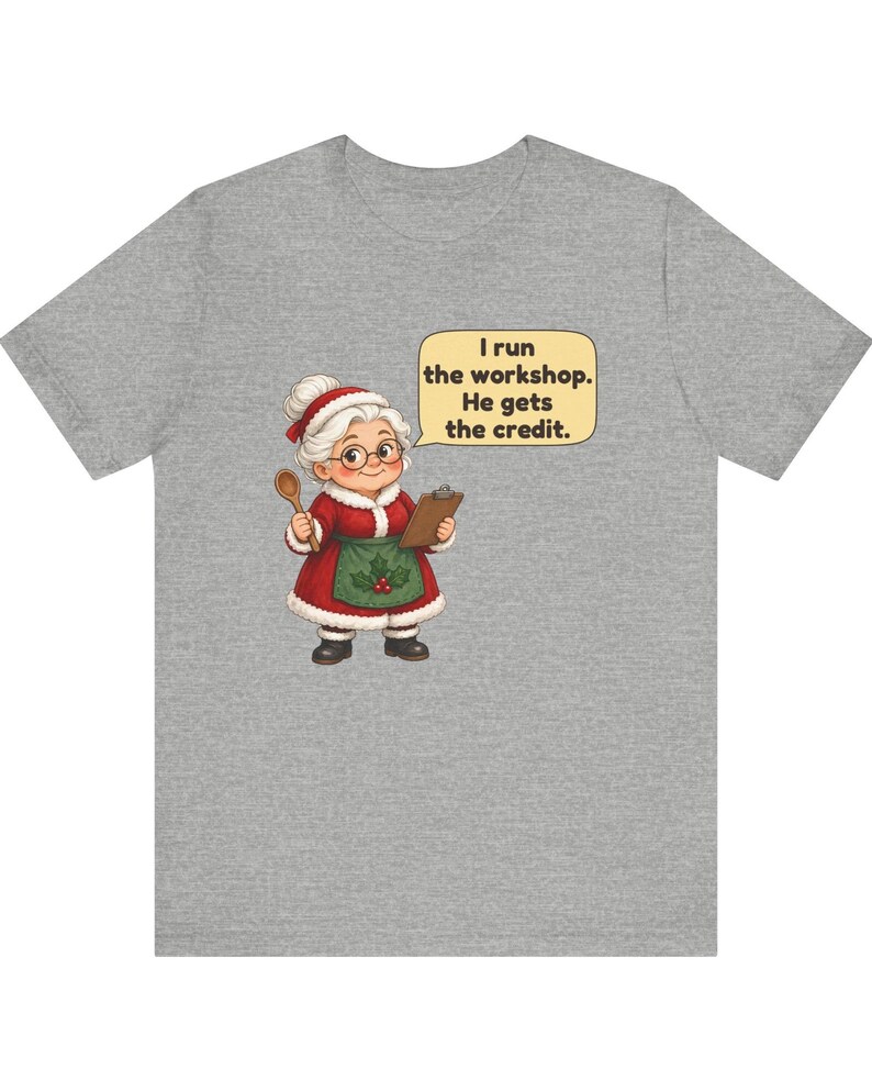 K&ouml;nnte beinhalten: Ein graumeliertes T-Shirt mit einer Cartoon-Illustration einer &auml;lteren Frau in einem rot-wei&szlig;en Festtagsoutfit, die einen Holzl&ouml;ffel und ein Klemmbrett h&auml;lt. Eine Sprechblase enth&auml;lt den Text: I run the workshop. He gets the credit.