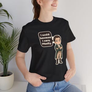 May include: A black short-sleeved t-shirt featuring a cartoon illustration of a person in a dark green coat and tan trousers with arms crossed. A speech bubble next to the character contains the text: I scold because I care. Mostly.