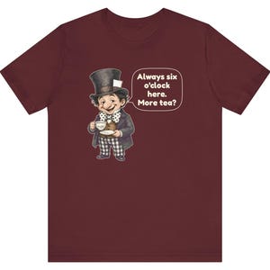 May include: A maroon t-shirt featuring a graphic of a character wearing a top hat, a polka-dot bow tie, and checkered trousers, holding a teacup and saucer. A speech bubble next to the character contains the text: Always six o'clock here. More tea?