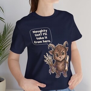 Puede incluir: Una camiseta azul marino con el gr&aacute;fico de una criatura marr&oacute;n con cuernos que sostiene un manojo de ramas y un pergamino. Un globo de texto sobre la criatura contiene el texto: Naughty list? I'll take it from here.