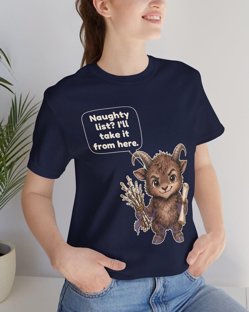 Puede incluir: Una camiseta azul marino con el gr&aacute;fico de una criatura marr&oacute;n con cuernos que sostiene un manojo de ramas y un pergamino. Un globo de texto sobre la criatura contiene el texto: Naughty list? I'll take it from here.
