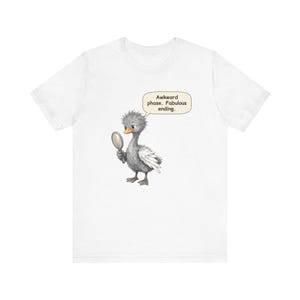 May include: A white t-shirt featuring a graphic of a fluffy gray duckling holding a small hand mirror. A speech bubble above the duckling contains the text: Awkward phase. Fabulous ending. The duckling has a bright orange beak and white wing feathers.
