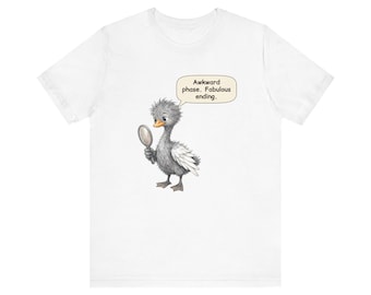 May include: A white t-shirt featuring a graphic of a fluffy gray duckling holding a small hand mirror. A speech bubble above the duckling contains the text: Awkward phase. Fabulous ending. The duckling has a bright orange beak and white wing feathers.