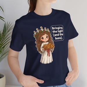 May include: A navy blue t-shirt featuring a cartoon illustration of a figure wearing a white robe and a candle crown, holding a tray of buns. The graphic includes a speech bubble with the text: Bringing the light (and the buns).