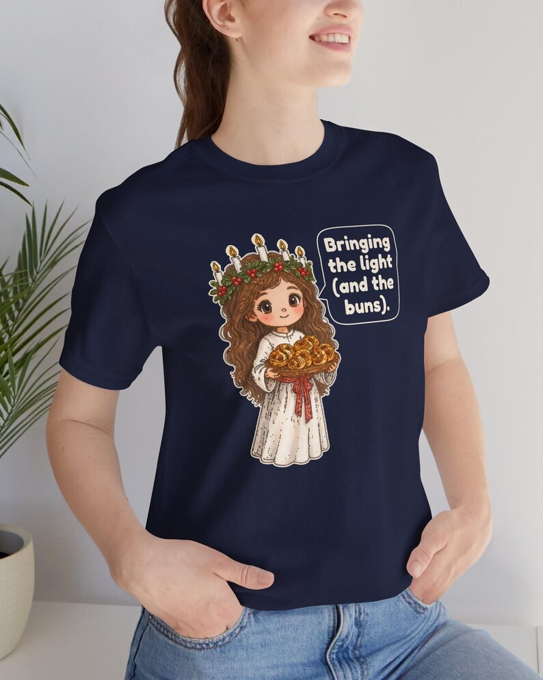 May include: A navy blue t-shirt featuring a cartoon illustration of a figure wearing a white robe and a candle crown, holding a tray of buns. The graphic includes a speech bubble with the text: Bringing the light (and the buns).