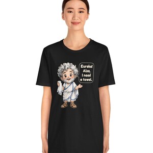 May include: A black t-shirt featuring a cartoon illustration of a bearded man in a white toga holding a white cloth. A speech bubble next to the figure contains the text: Eureka! Also, I need a towel. The design is centered on the front of the shirt.