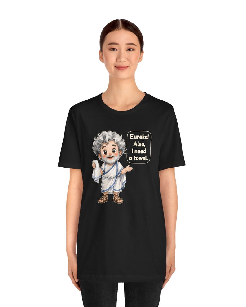 May include: A black t-shirt featuring a cartoon illustration of a bearded man in a white toga holding a white cloth. A speech bubble next to the figure contains the text: Eureka! Also, I need a towel. The design is centered on the front of the shirt.