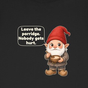 Op de afbeelding: Een zwart t-shirt met een afbeelding van een kabouter met een rode muts, die een houten kom met pap en een lepel vasthoudt. Een tekstballon naast de kabouter bevat de tekst: Leave the porridge. Nobody gets hurt.