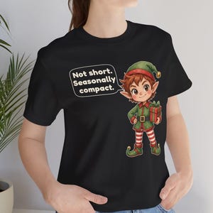 May include: A black t-shirt featuring a cartoon elf wearing a green hat and outfit with red and white striped leggings, holding a red gift box. A speech bubble next to the elf contains the text: Not short. Seasonally compact.