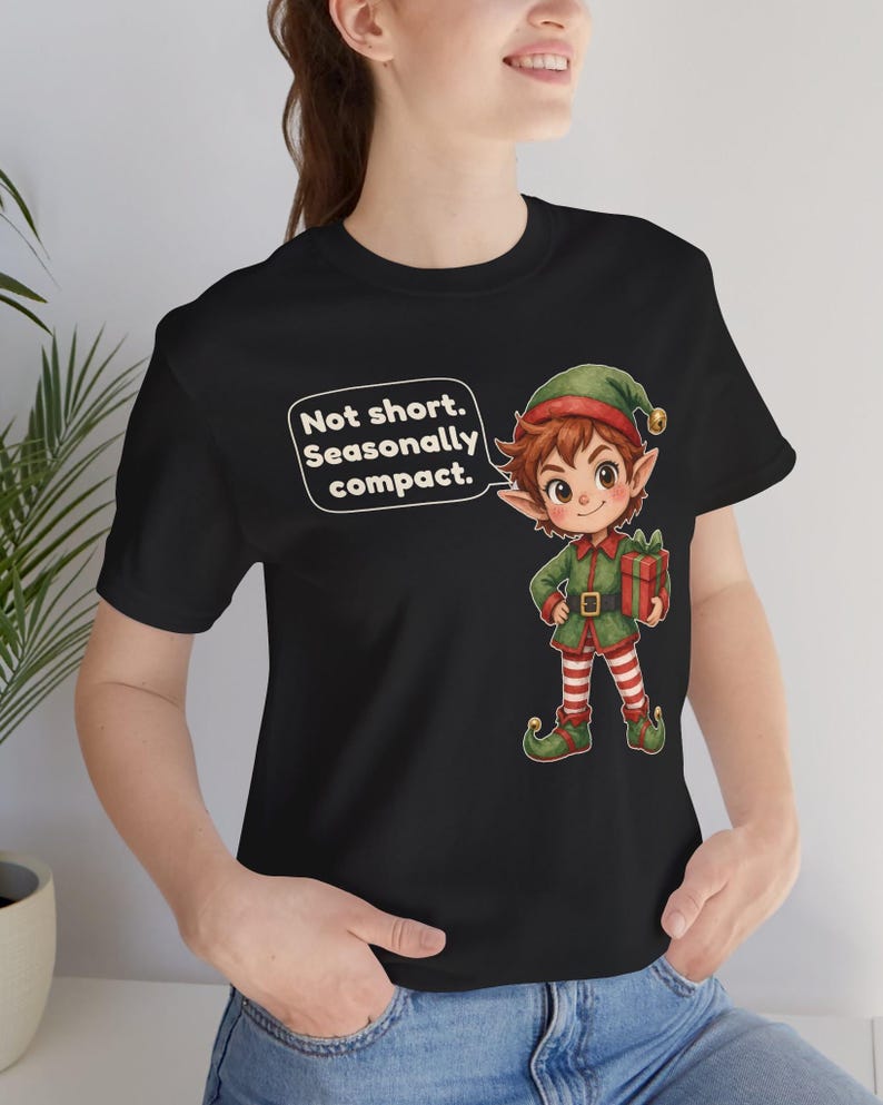 May include: A black t-shirt featuring a cartoon elf wearing a green hat and outfit with red and white striped leggings, holding a red gift box. A speech bubble next to the elf contains the text: Not short. Seasonally compact.