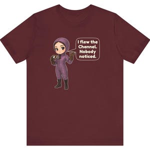 Puede incluir: Una camiseta de manga corta color granate con un personaje de dibujos animados con traje de vuelo morado y gafas. Un bocadillo junto al personaje contiene el texto en blanco: I flew the Channel. Nobody noticed.