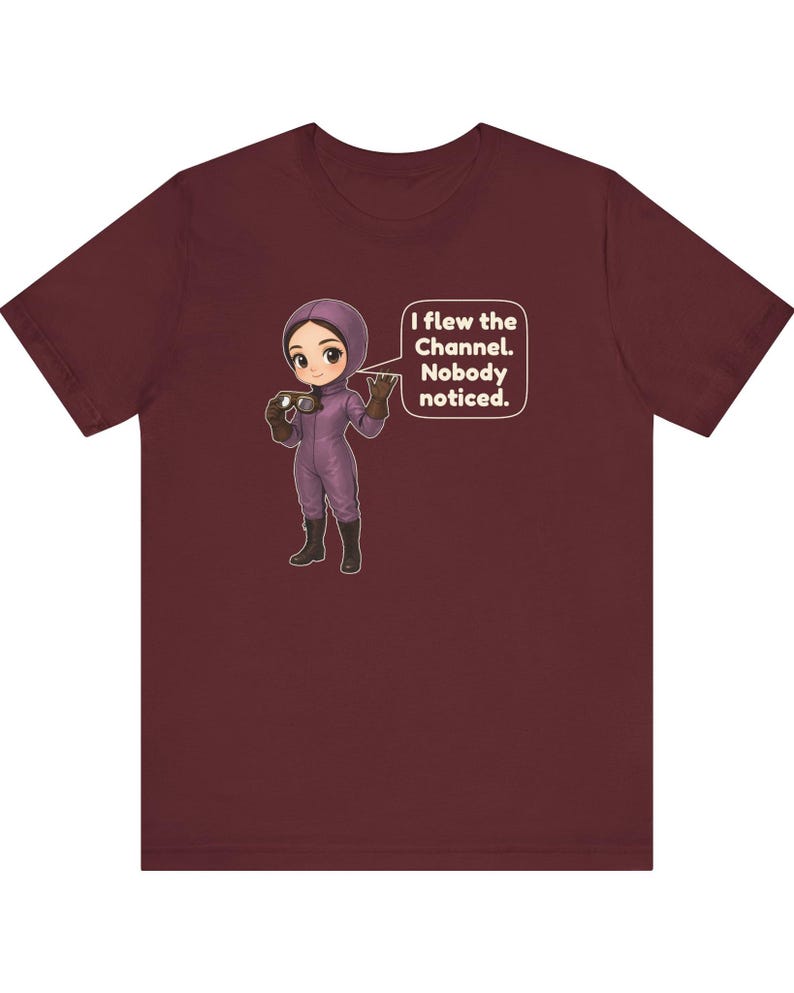 Puede incluir: Una camiseta de manga corta color granate con un personaje de dibujos animados con traje de vuelo morado y gafas. Un bocadillo junto al personaje contiene el texto en blanco: I flew the Channel. Nobody noticed.
