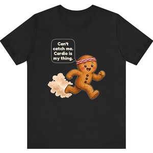May include: A black t-shirt featuring a cartoon gingerbread person wearing a red and white striped headband, running and leaving a dust cloud behind. A speech bubble above the character contains the text: Can't catch me. Cardio is my thing.