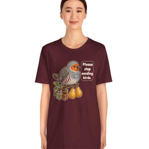 K&ouml;nnte beinhalten: Ein kastanienbraunes Kurzarm-T-Shirt mit einer Grafik eines Rotkehlchens auf einem Ast, zusammen mit zwei Birnen und zwei kleinen Geschenken. Der Vogel steht neben einer Sprechblase mit dem Text: "Please stop sending birds."