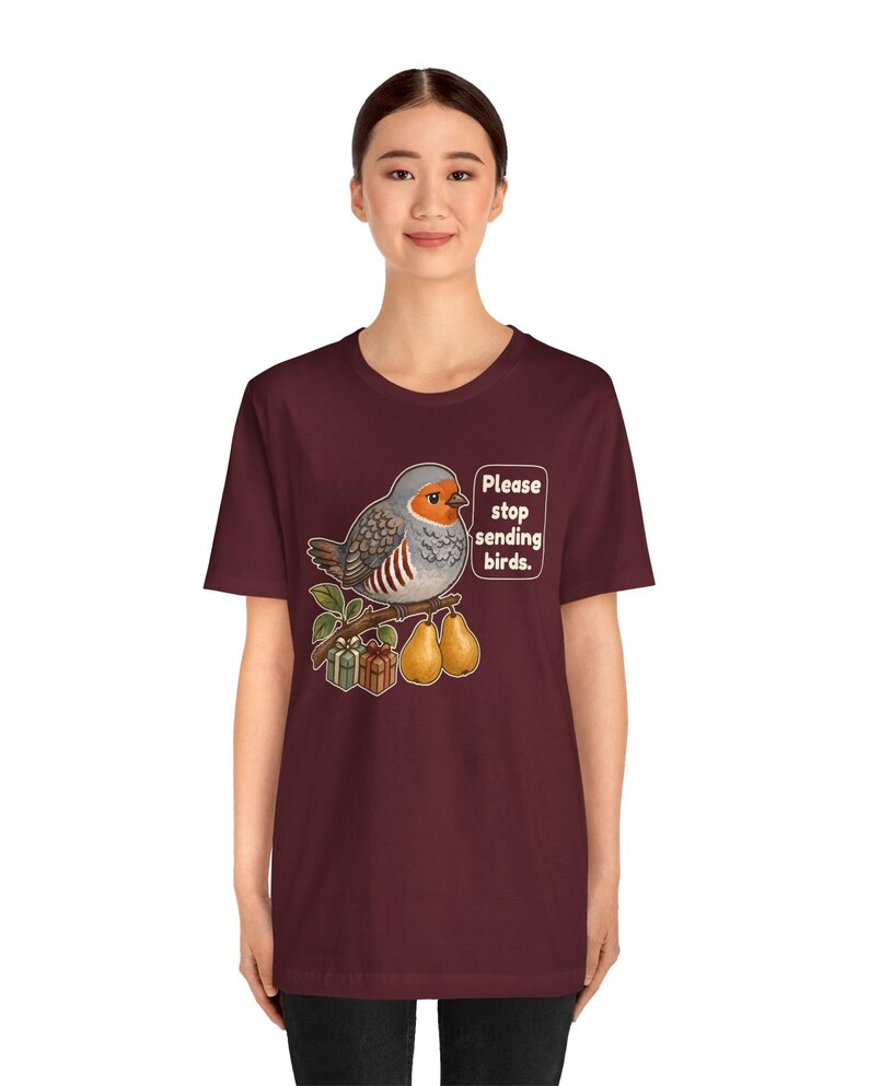 K&ouml;nnte beinhalten: Ein kastanienbraunes Kurzarm-T-Shirt mit einer Grafik eines Rotkehlchens auf einem Ast, zusammen mit zwei Birnen und zwei kleinen Geschenken. Der Vogel steht neben einer Sprechblase mit dem Text: "Please stop sending birds."
