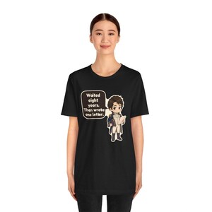 Puede incluir: Una camiseta negra de manga corta con un personaje de dibujos animados con uniforme militar azul y blanco que sostiene una carta. Un globo de texto junto al personaje contiene el texto: Waited eight years. Then wrote one letter.